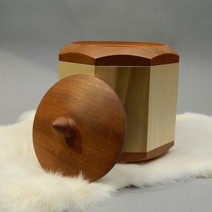 May include: A wooden box with a lid, featuring a geometric design. The box is made of light and dark wood, with a lid that is partially open. The box is sitting on a white, fluffy surface.