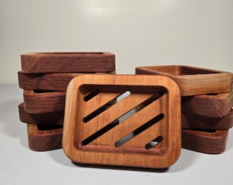 May include: Wooden soap dishes in various shades of brown. The front-most dish has a rectangular shape with diagonal slots. Several other dishes are stacked behind it. The wood grain is visible on the surface.
