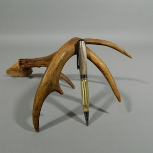 May include: A brown and black pen with a brass body and a black clip is displayed on a set of antlers. The pen is made of wood and has a unique design.