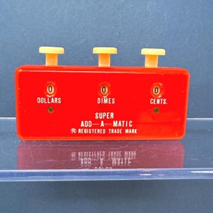 May include: A bright red rectangular Super Add-A-Matic mechanical adding device with three cream-colored push buttons on top. The front features three numerical display windows labeled Dollars, Dimes, and Cents, with white printed text throughout.