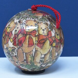 May include: A round Christmas ornament featuring a repeating pattern of teddy bears with red bows and green foliage. The ornament has a red cord for hanging. The background is a gradient of white and blue.