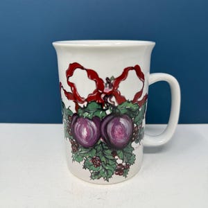 May include: White ceramic mug with a Christmas-themed design. The design features a red bow, holly leaves, and purple berries.