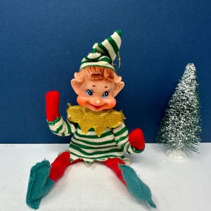 May include: A vintage Christmas elf doll with a green and white striped hat and shirt, red gloves and pants, and teal shoes. The elf has a gold collar and is posed with a small, silver Christmas tree in the background.