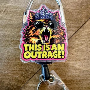 May include: A retractable badge reel with a cartoon cat wearing sunglasses and a crown. The cat has an open mouth and is surrounded by orange, pink, and black fur. The text "THIS IS AN OUTRAGE!" is in yellow and pink.