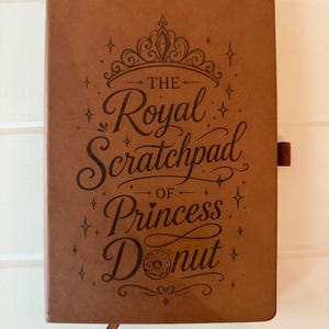 May include: A tan leather-like notebook featuring a dark brown laser-engraved design of a tiara and the text "THE Royal Scratchpad OF Princess Donut" with a small donut illustration. The notebook includes a brown elastic pen loop on the side.