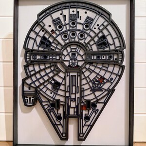 May include: A multi-layered, laser-cut wooden wall art piece depicting the Millennium Falcon spaceship. The design features intricate black geometric cutouts layered over a white background, all enclosed within a rectangular black frame.