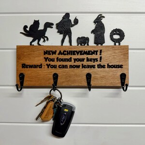 May include: A rectangular wooden key holder with four black metal hooks. The board features the text: NEW ACHIEVEMENT ! You found your keys ! Reward : You can now leave the house. Above the text are five black silhouette figures.