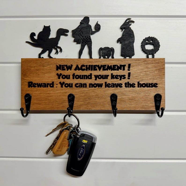 May include: A rectangular wooden key holder with four black metal hooks. The board features the text: NEW ACHIEVEMENT ! You found your keys ! Reward : You can now leave the house. Above the text are five black silhouette figures.
