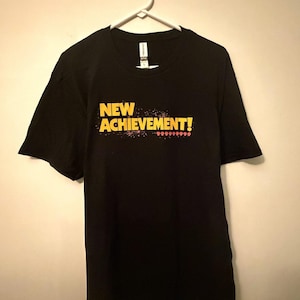 May include: A black short-sleeve t-shirt featuring the yellow text NEW ACHIEVEMENT! printed on the chest. Below the text is a row of small red pixelated skull icons. The shirt is displayed on a white plastic hanger against a plain wall.