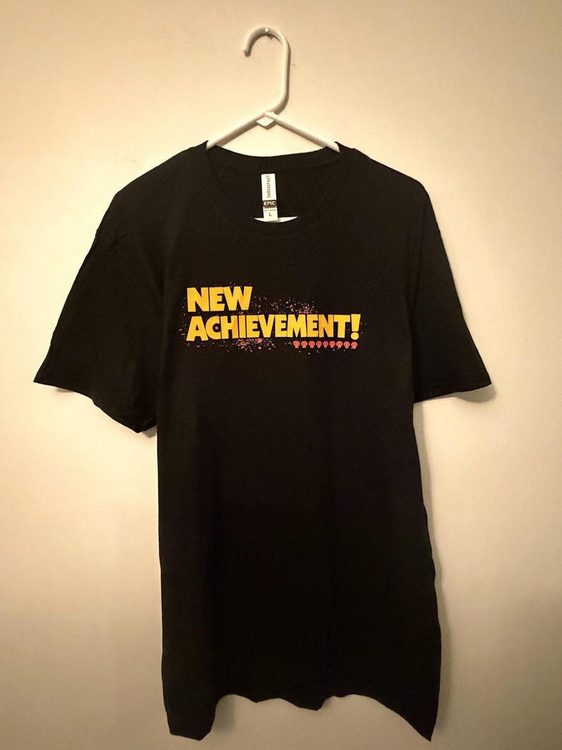 May include: A black short-sleeve t-shirt featuring the yellow text NEW ACHIEVEMENT! printed on the chest. Below the text is a row of small red pixelated skull icons. The shirt is displayed on a white plastic hanger against a plain wall.