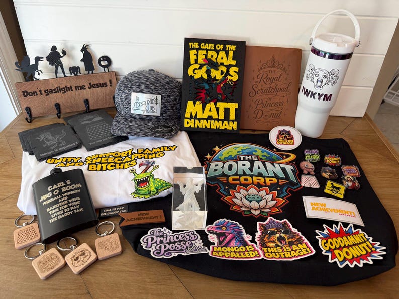 May include: A collection of merchandise including a black hat, a book titled The Gate of the Feral Gods, a white water bottle with the text IWKYM, a black flask labeled Carl's Jug O Boom, a white t-shirt, a black tote bag, and various stickers.