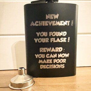 May include: A matte black metal flask with a screw-top cap and a silver-colored metal funnel. The flask is engraved with the text: NEW ACHIEVEMENT! YOU FOUND YOUR FLASK! REWARD: YOU CAN NOW MAKE POOR DECISIONS. The design is simple and functional.