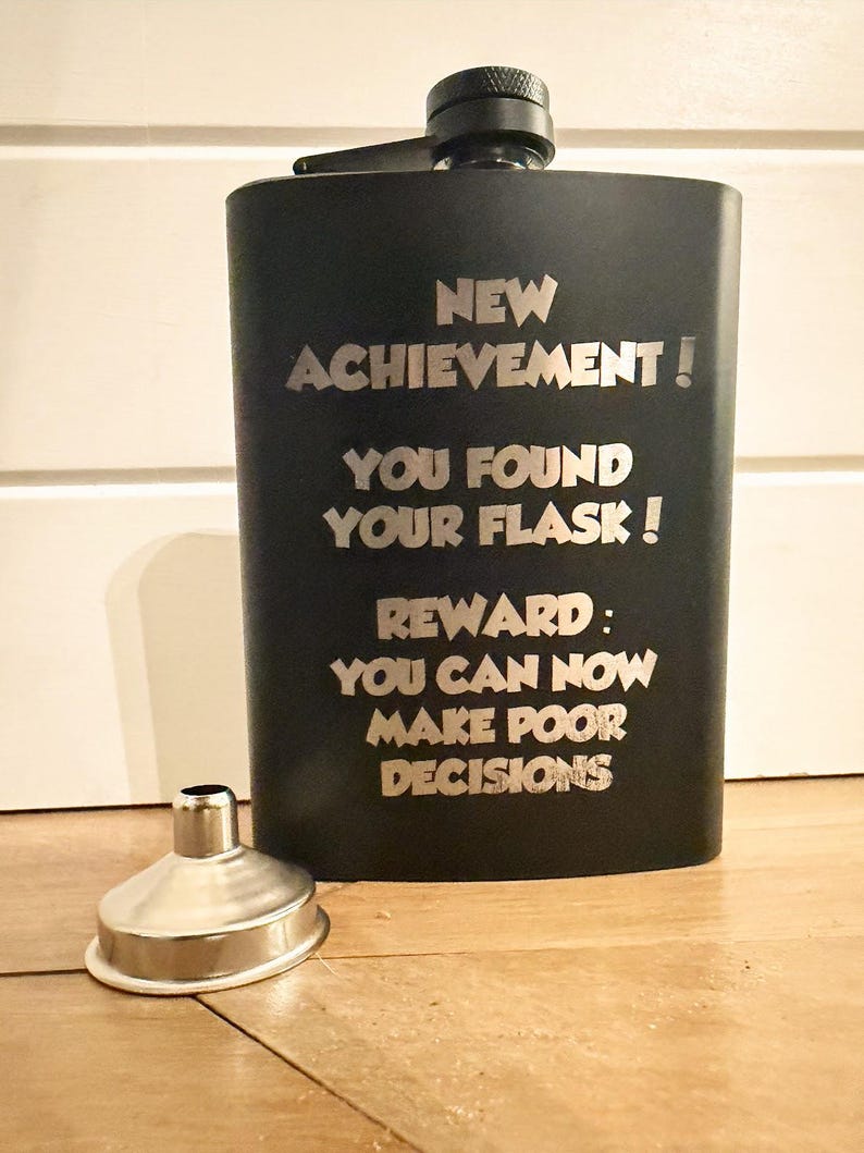 May include: A matte black metal flask with a screw-top cap and a silver-colored metal funnel. The flask is engraved with the text: NEW ACHIEVEMENT! YOU FOUND YOUR FLASK! REWARD: YOU CAN NOW MAKE POOR DECISIONS. The design is simple and functional.