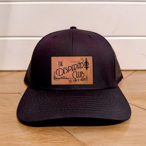 May include: Black trucker hat with a curved brim and mesh back. The front features a rectangular patch with the words "The Desperado Club" and the phrase "So Fun It Hurts!" in a stylized font. The patch has a brown leather-like appearance.