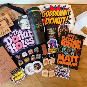 May include: A collection of merchandise including a black tote bag with The Donut Holes text, a black tumbler, a white t-shirt with Goddamnit Donut text, a white baseball cap, a book titled The Eye of the Bedlam Bride, stickers, keychains, and coasters.