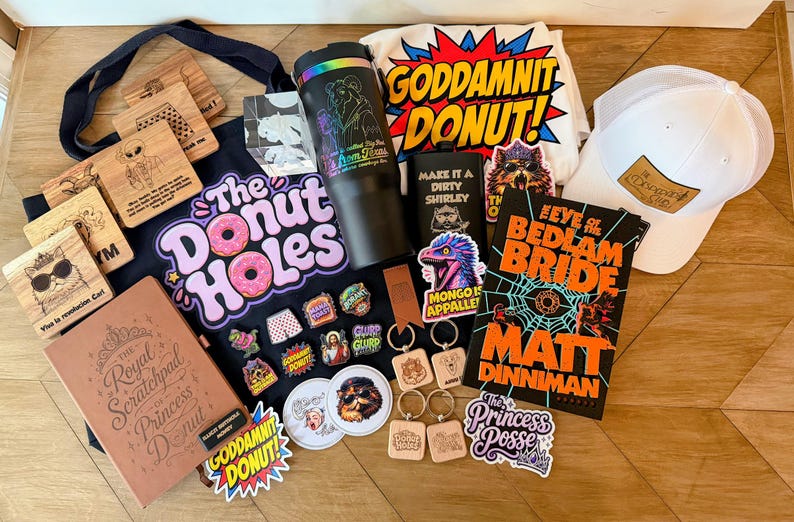 May include: A collection of merchandise including a black tote bag with The Donut Holes text, a black tumbler, a white t-shirt with Goddamnit Donut text, a white baseball cap, a book titled The Eye of the Bedlam Bride, stickers, keychains, and coasters.