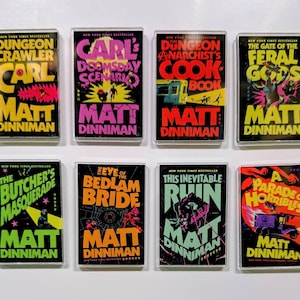 May include: A collection of eight rectangular magnets featuring book covers by Matt Dinniman. The covers have vibrant colors and bold text, with titles like "Dungeon Crawler" and "The Butcher's Masquerade". Each magnet is encased in a clear plastic frame.