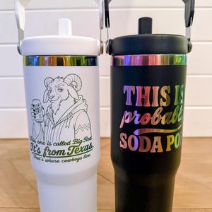 May include: A white tumbler with a white handle and a black tumbler with a black handle. The white tumbler features a goat illustration holding a Big Red can with text reading This one is called Big Red. It's from Texas. That's where cowboys live. The black tumbler has iridescent text reading THIS IS probably SODA POP.