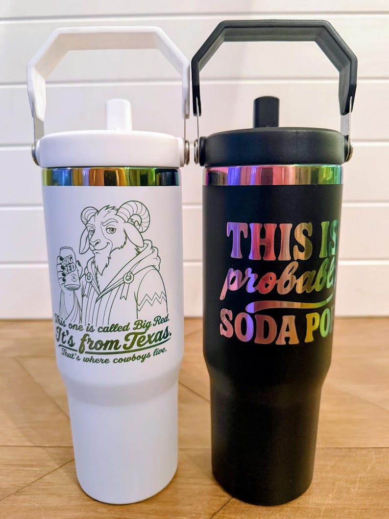 May include: A white tumbler with a white handle and a black tumbler with a black handle. The white tumbler features a goat illustration holding a Big Red can with text reading This one is called Big Red. It's from Texas. That's where cowboys live. The black tumbler has iridescent text reading THIS IS probably SODA POP.