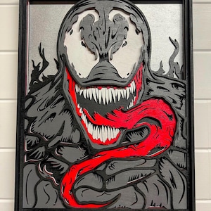 May include: Layered wood art of the Marvel Comics character Venom. The artwork features a black frame, with a gray and black Venom face, white teeth, and a red tongue. The background is a light gray color.