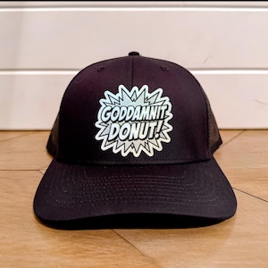 May include: Black trucker hat with a mesh back and a curved brim. The front of the hat features a white and blue graphic that reads "GODDAMNIT DONUT!" in a comic book style font.