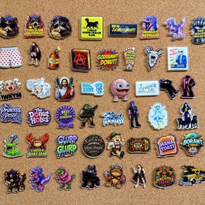 May include: A collection of assorted enamel pins featuring various graphic designs, including cartoon characters, animals, text-based slogans, and pop culture references in diverse shapes, colors, and sizes arranged on a cork surface.