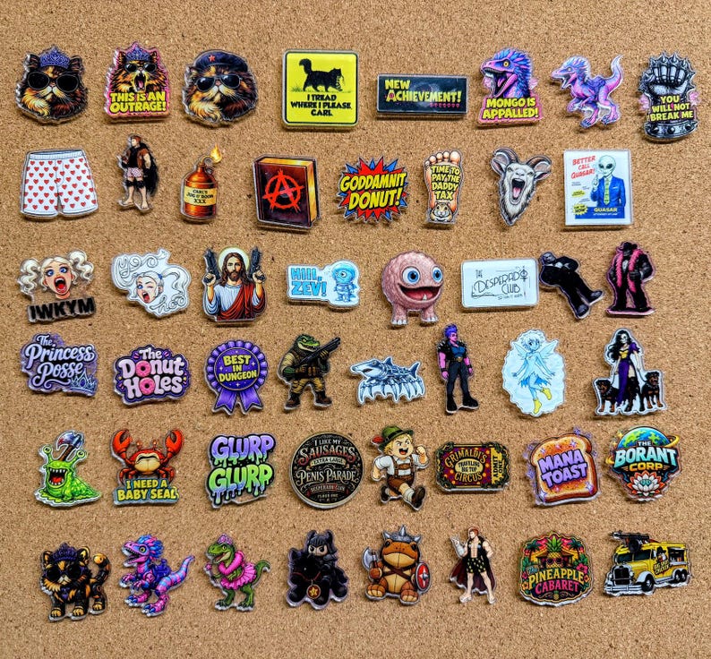 May include: A collection of assorted enamel pins featuring various graphic designs, including cartoon characters, animals, text-based slogans, and pop culture references in diverse shapes, colors, and sizes arranged on a cork surface.