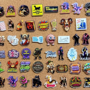 May include: A collection of assorted enamel pins featuring various graphic designs, including cartoon characters, animals, text-based slogans, and pop culture references in vibrant colors, arranged in a grid on a textured cork surface.