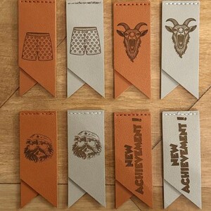 May include: Eight rectangular leather tags in tan and light gray, featuring stitched tops and pointed bottoms. Designs include boxer shorts with hearts, a goat head, a bearded character with a beret and glasses, and the text NEW ACHIEVEMENT!