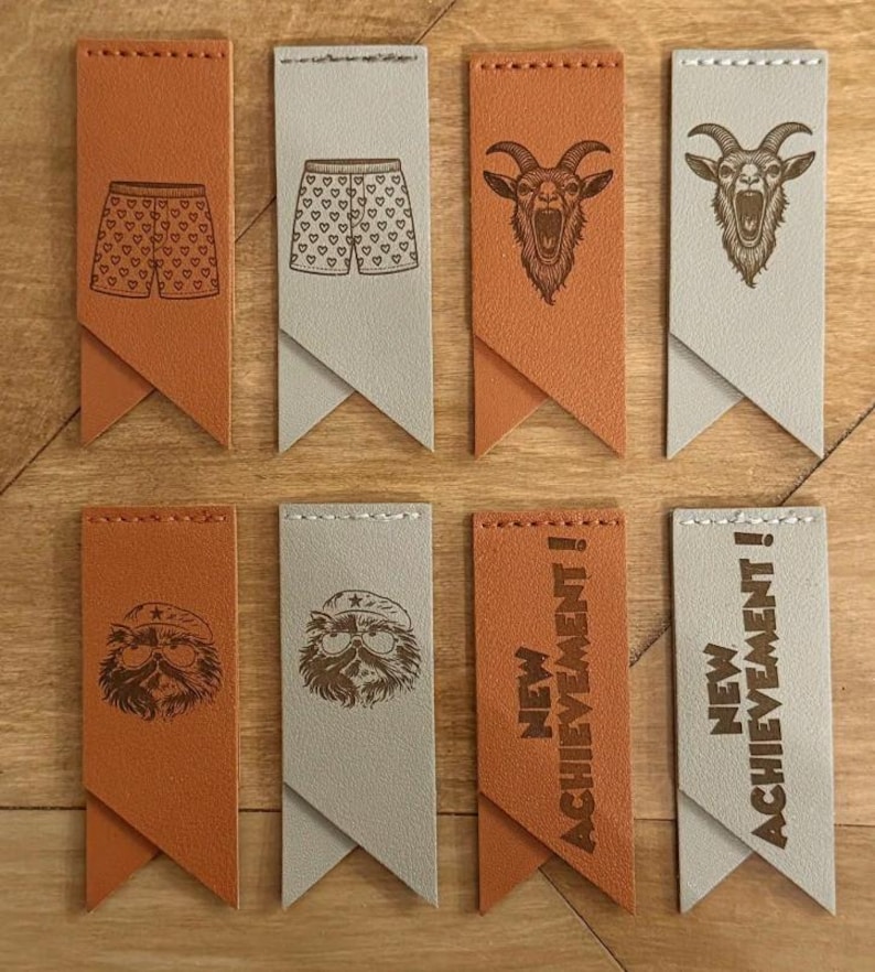 May include: Eight rectangular leather tags in tan and light gray, featuring stitched tops and pointed bottoms. Designs include boxer shorts with hearts, a goat head, a bearded character with a beret and glasses, and the text NEW ACHIEVEMENT!