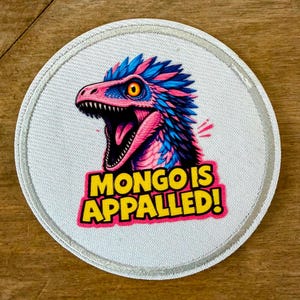 May include: Round patch with a colorful dinosaur illustration. The dinosaur has pink, blue, and yellow accents, with the text "MONGO IS APPALLED!" in a yellow and pink font. The patch has a white background and a silver border.
