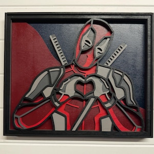 May include: A framed, layered wood art piece featuring the Deadpool character. The artwork uses red, gray, and black colors to depict Deadpool making a heart shape with his hands. The background is a mix of red and blue.