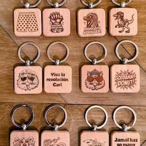 May include: A collection of twelve square wooden keychains with silver metal rings. Each keychain features a unique laser-engraved design, including illustrations of a dinosaur, a cat with sunglasses, a shark, and various text phrases like "Viva la revoluci&oacute;n, Carl."