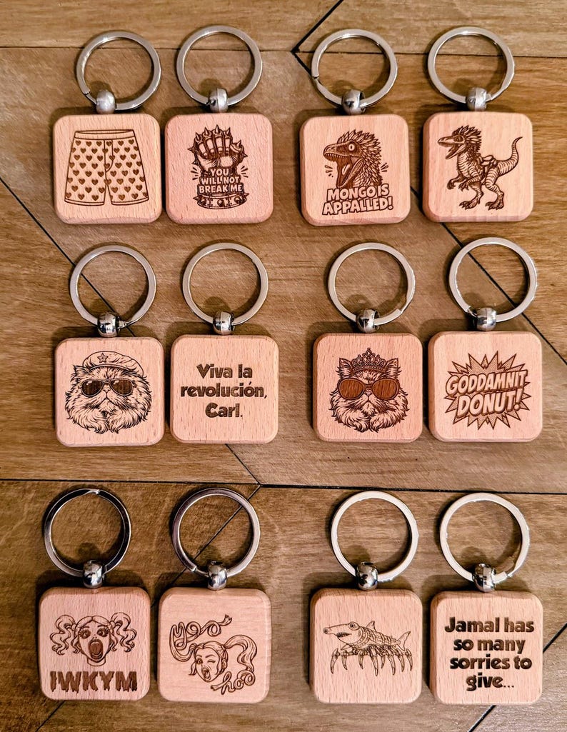 May include: A collection of twelve square wooden keychains with silver metal rings. Each keychain features a unique laser-engraved design, including illustrations of a dinosaur, a cat with sunglasses, a shark, and various text phrases like "Viva la revoluci&oacute;n, Carl."