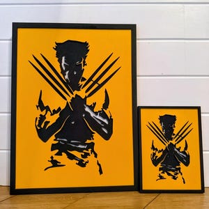 May include: Two framed art prints featuring a black silhouette of a superhero with claws extended. The artwork is set against a bright yellow background and framed in black. The larger print is rectangular, and the smaller one is square.