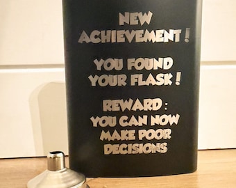 May include: A black flask with a silver screw-on cap and a silver funnel. The flask has the text "NEW ACHIEVEMENT! YOU FOUND YOUR FLASK! REWARD: YOU CAN NOW MAKE POOR DECISIONS" in silver lettering.