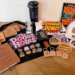 May include: A collection of merchandise including a black tote bag with The Donut Holes text, a brown notebook, a black tumbler, a white t-shirt with Goddamnit Donut text, a white baseball cap, a book, various enamel pins, keychains, and stickers.