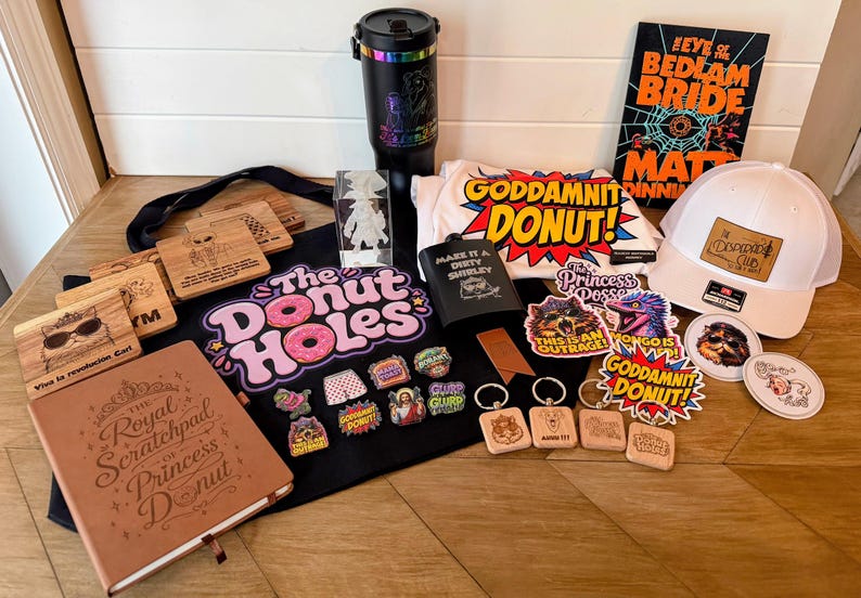 May include: A collection of merchandise including a black tote bag with The Donut Holes text, a brown notebook, a black tumbler, a white t-shirt with Goddamnit Donut text, a white baseball cap, a book, various enamel pins, keychains, and stickers.
