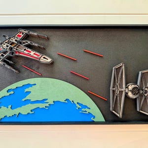 May include: A framed wooden artwork featuring a red and white X-wing fighter and a black and silver TIE fighter. The scene includes red laser blasts and a green and blue planet against a black background.