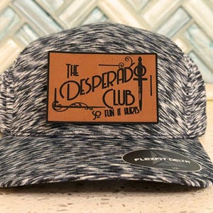 May include: A black and white heathered baseball cap featuring a rectangular tan leather patch on the front. The patch displays the text "THE DESPERADO CLUB" and "SO FUN IT HURTS" with a sword graphic. A black Flexfit Delta sticker is on the brim.