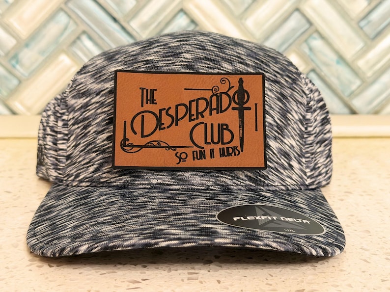 May include: A black and white heathered baseball cap featuring a rectangular tan leather patch on the front. The patch displays the text "THE DESPERADO CLUB" and "SO FUN IT HURTS" with a sword graphic. A black Flexfit Delta sticker is on the brim.