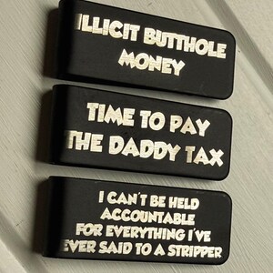 May include: Three rectangular black metal money clips featuring laser-engraved text. The top clip reads ILLICIT BUTTHOLE MONEY, the middle clip reads TIME TO PAY THE DADDY TAX, and the bottom clip reads I CAN'T BE HELD ACCOUNTABLE FOR EVERYTHING I'VE EVER SAID TO A STRIPPER.