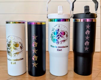 May include: Four cylindrical tumblers in black and white with iridescent accents. Two feature a cat wearing a beret and sunglasses with the text "Viva la revolución Carl." The other two feature a vertical column of five star and anarchy symbols.