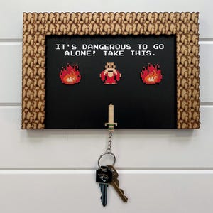 Op de afbeelding: Een houten sleutelhouder met een zwarte achtergrond en een pixelontwerp. De tekst "IT'S DANGEROUS TO GO ALONE! TAKE THIS." wordt weergegeven boven gepixelde vlammen en een personage. Sleutels hangen aan een zwaardvormige sleutelhanger.