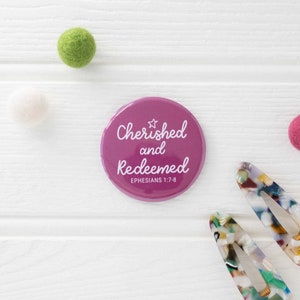 May include: A pink button with the text "Cherished and Redeemed" and the scripture reference "Ephesians 1:7-8" in white lettering.