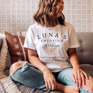 May include: A white t-shirt with the text "LUNA'S EMPORIUM of Oddities" and a small illustration of a turnip. The text below the logo reads "For the Exceptionally Ordinary Witch and Wizard."