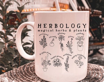 May include: A white ceramic mug with the word "HERBOLOGY" and the phrase "magical herbs & plants" printed in black. Below are illustrations of various herbs and plants, including wormwood and mandrake.