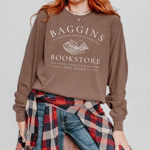 May include: Brown long-sleeve shirt with a white graphic design that says "Baggins Bookstore" with a book illustration and the text "An unexpected journey on every page. Open everyday after second breakfast - The Shire."