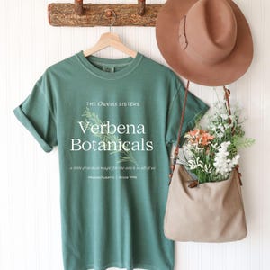 Puede incluir: Una camiseta verde con el texto "Verbena Botanicals" y una ilustración botánica. La camiseta está colgada en una percha de madera. También se ven un sombrero marrón y un bolso beige lleno de flores.