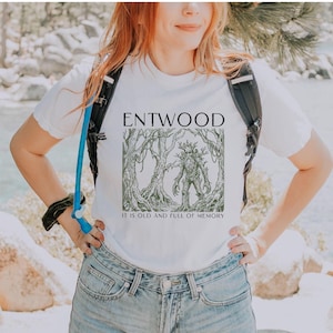 以下が含まれることがあります： 「ENTWOOD」の文字と、木のような姿の人物が描かれた森のイラストがプリントされた白いTシャツ。イラストの下には「IT IS OLD AND FULL OF MEMORY」の文字。青いデニムのショートパンツと合わせて着用。